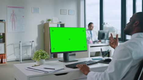 African-American-Medical-Doctor-macht-einen-Videoanruf-mit-Patient-auf-einem-Computer-mit-Green-Screen-Display-in-einer-Gesundheitsklinik.-Assistant-in-Lab-Coat-spricht-über-Gesundheitsprobleme-im-Krankenhausbüro.