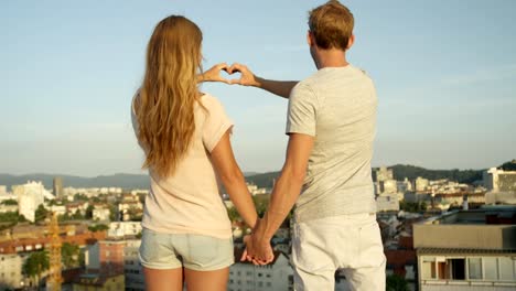 CLOSE-UP:-Couple-standing-on-rooftop-holding-hands-making-heart-with-fingers