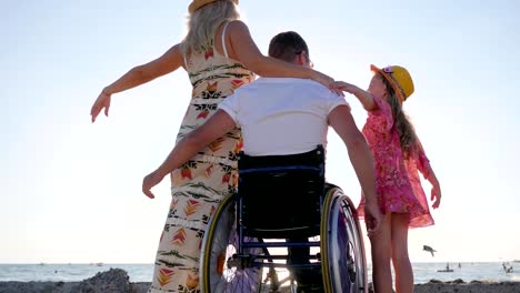 familia-feliz-levantando-manos-mirando-las-olas,-vista-trasera,-esposo-esposa-abrazo-de-silla-de-ruedas-y-niña,-abrazos-con-discapacidad