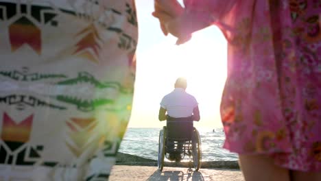 mother-and-daughter-keep-hands-against-backdrop-of-father-sitting-in-wheelchair