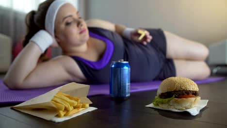 Faul-Dame-in-Sportbekleidung-liegen-auf-Yoga-Matte-Essen-Junk-Food-statt-training