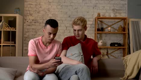 Two-international-friends-of-a-homosexual-are-sitting-on-the-couch-and-watching-disgusting,-mercenary-pictures-on-a-smartphone.-Home-cosiness,-family,-internet-concept.-60-fps