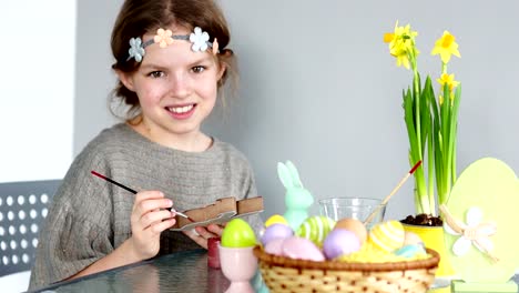 A-brown-eyed-girl-with-a-wreath-on-her-head-is-preparing-for-Easter.-The-child-looks-at-the-camera-and-laughs-cheerfully.-In-her-hands-paint-and-brush.-On-the-table-is-an-Easter-basket-and-a-bouquet-of-daffodils