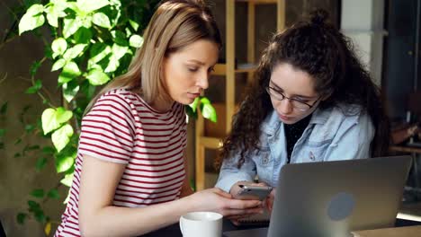 Close-up-of-two-female-students-using-smartphone.-Attractive-blond-girl-is-pointing-touchscreen-and-chatting-with-her-friend.-Modern-technology-for-young-people-concept.