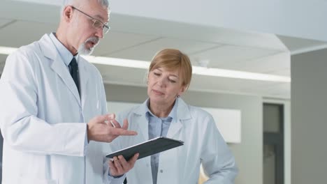 In-the-Hospital-Doctors-have-Discussion-while-Using-Tablet-Computer.-In-the-Background-Patients-and-Medical-Personnel.-New-Modern-Fully-Functional-Medical-Facility.