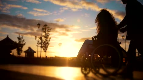 Caring-man-with-a-disabled-woman-in-wheelchair-walking-through-the-quay-at-sunset