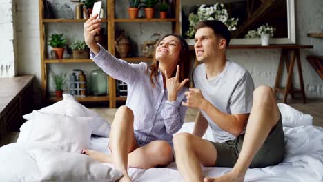 Modern-married-couple-is-taking-selfie-in-bedroom-gesturing-posing-and-kissing-while-sitting-on-bed-together.-Modern-technology-and-people-concept.