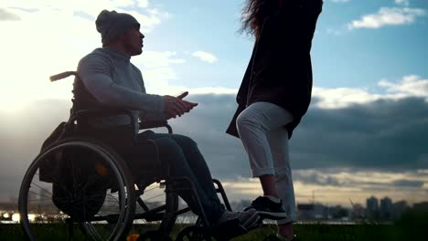 Young-woman-with-disabled-man-in-a-wheelchair-talking-outdoors-at-sunset