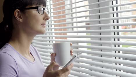 Junge-Frau-in-Gläsern-trinkt-Kaffee-stehen-mit-Smartphone-durch-Fenster-im-Büro.-Mädchen-schaut-aus-dem-Fenster-und-nutzt-eine-Smartphone.-SMS-Nachricht,-online-surfen,-app,-Soziales-Netzwerk.