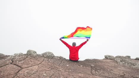 Mann-erhöhen-Regenbogen-Farbe-LGBTI-Fahnenschwingen-in-harten-Wind-auf-Mountain-Top-Sicht