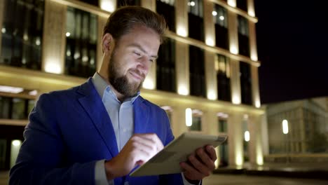 Panning-Medium-shot-der-schönen-bärtigen-Geschäftsmann-in-blaue-Jacke-mit-Tablet-Computer-in-der-Nacht-beleuchtete-Stadt-Straße