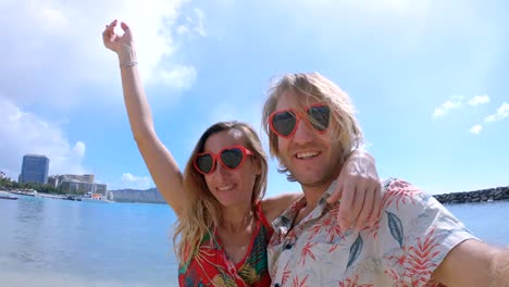 Couple-taking-selfies-on-Waikiki-Beach-in-Honolulu,-Hawaii.-Selfie-point-of-view-wide-angle-of-Waikiki-Beach.-Young-couple-taking-selfies-with-heart-shaped-sunglasses