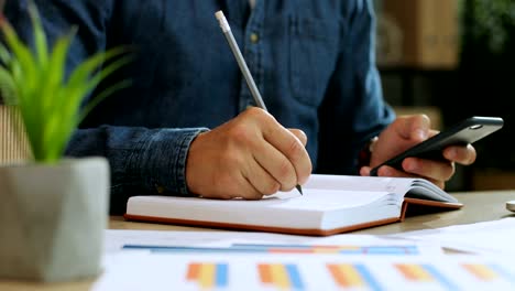 Cerca-de-un-tiro-de-manos-de-hombre-joven-usando-el-teléfono-inteligente-fot-señalando-algo-en-el-libro-de-notas-mientras-trabajaba-en-la-oficina.