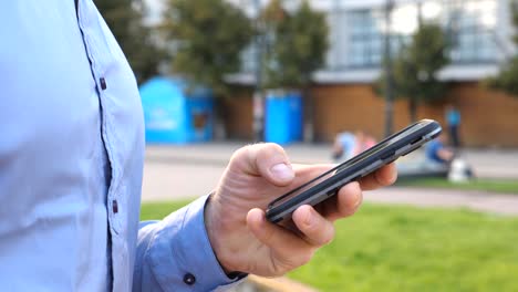 Cerrar-mano-de-joven-empresario-sosteniendo-y-tocando-el-teléfono-inteligente.-Hombre-irreconocible-parado-en-la-calle-urbana-y-noticias-de-navegación-en-el-teléfono.-Un-tipo-usando-artilugio-para-trabajar.-La-cámara-lenta-Dolly-disparó