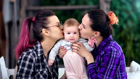 Cute-little-baby-posing-looking-at-camera-during-two-happy-mother-kissing-and-hugging-him