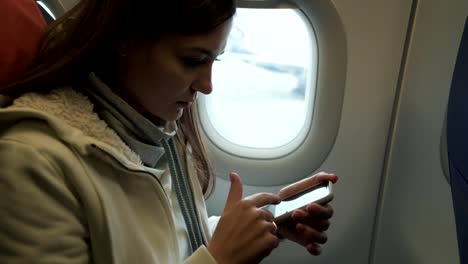 Portrait-young-girl-travels-by-plane,-sits-near-window-and-uses-phone