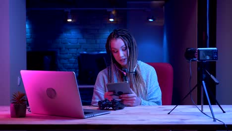 Closeup-shoot-of-young-attractive-female-blogger-with-dreadlocks-in-headphones-streaming-live-using-the-tablet-with-the-neon-background-indoors