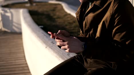 Side-view-of-a-elegant-nab-in-glasses-and-brown-coat-sitting-outside-on-parapet-near-the-seaside.-Brunette-young-man-looking-at-his-smarphone,-texting.-Sunny,-windy-cool-weather