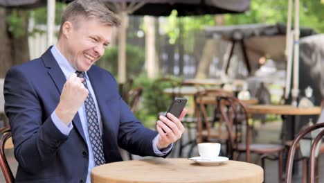 Empresario-celebrando-el-éxito-en-el-teléfono,-sentado-en-el-café-al-aire-libre