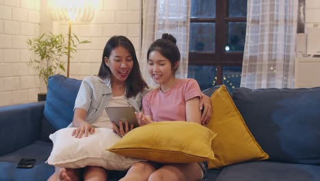 Lesben-lgbt-Frauen-Paar-mit-Tablet-zu-Hause,-asiatische-Frau-glücklich-mit-Technologie-überprüfen-Social-Media-im-Internet-zusammen,-während-liegen-Sofa-im-Wohnzimmer-in-der-Nacht.-Liebhaber-feiern-Urlaubskonzept.