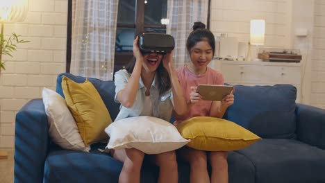 Lesbianas-mujeres-lgbt-pareja-usando-tableta-en-casa,-mujer-asiática-se-siente-feliz-usando-computadora-portátil-y-VR-jugando-juegos-juntos-mientras-se-acuesta-sofá-en-la-sala-de-estar-en-la-noche.-Los-amantes-celebran-el-concepto-de-vacaciones.