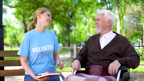 Young-female-volunteer-reading-book-for-disabled-pensioner-in-wheelchair,-care