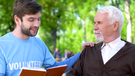 Amable-libro-de-lectura-de-voluntarios-masculinos-a-un-anciano-sonriente-en-silla-de-ruedas,-asistencia