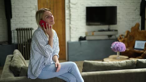 Mujer-caucásica-sonriente-de-25-años-con-ropa-casual-teniendo-conversación-de-teléfono-móvil-mientras-está-sentada-en-el-sofá