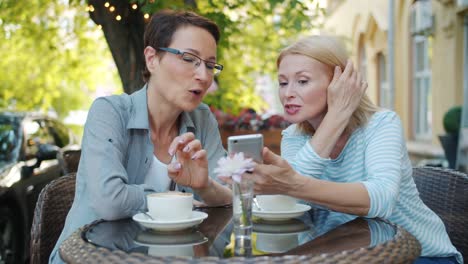 Cheerful-ladies-expressing-positive-emotions-using-smartphone-in-cafe-outdoors