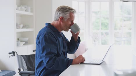 Anciano-discapacitado-hombre-en-silla-de-ruedas-en-casa-trabajando-en-el-ordenador-portátil-en-el-mostrador-de-la-cocina