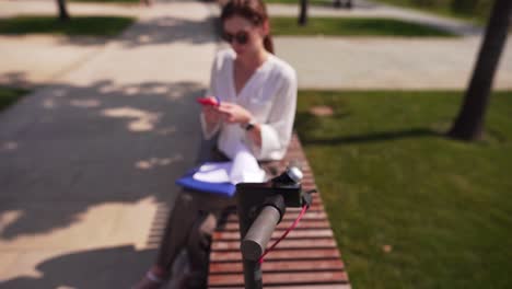 Female-hipster-with-a-smartphone-in-her-hands-makes-a-sale-via-the-Internet,-against-the-background-of-the-steering-wheel-from-an-electric-bike.