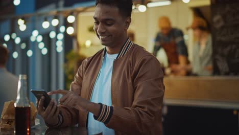 Handsome-Young-Indian-Man-está-usando-un-teléfono-inteligente-mientras-se-sienta-en-una-mesa-en-un-outdoors-Street-Food-Cafe.-Está-navegando-por-Internet-o-las-redes-sociales,-publicando-una-actualización-de-estado.-El-hombre-es-feliz-y-sonriente.