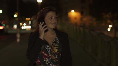 Glückliche-kaukasische-moderne-Frau-trägt-Blumenkleid,-schwarze-Jacke-und-rote-Haare-zu-Fuß-durch-die-Straße-und-telefonieren-auf-ihrem-Smartphone-in-der-Nacht.-Paris-4K-UHD.-Zeitlupe.