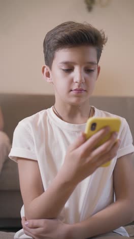 Portrait-of-Caucasian-brunette-boy-using-smart-phone.-Schoolchild-in-white-T-shirt-using-social-media-at-home.-Internet-addiction,-social-media-addiction.-Vertical-shooting.