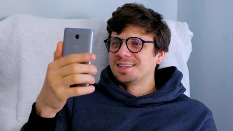 Joven-feliz-en-gafas-hablando-video-chat-en-el-teléfono-inteligente-sentado-en-el-sillón,-fecha-en-línea.