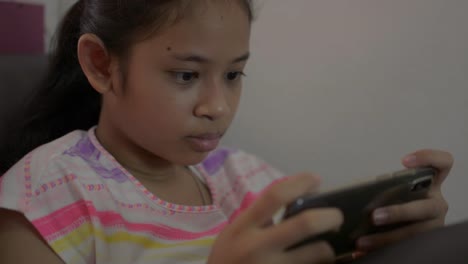 Adorable-girl-playing-games-on-mobile-smartphone-and-sitting-on-sofa-during-quarantine-at-home.-Asian-female-teenager-texting-on-social-online-communication-with-cell-phone.