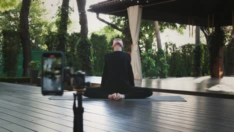 Calvo-hombre-con-ropa-deportiva-negra-está-realizando-yoga-sentado-en-esterilla-en-patio-ajardinado.-Disparándose-en-vídeo-usando-el-teléfono-inteligente-en-el-trípode.-De-cerca