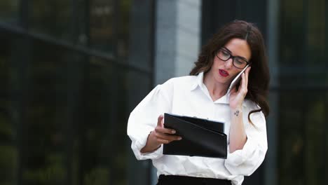 Mädchen-in-Brille-und-weißem-Hemd-Tanding-in-der-Nähe-von-Business-Center-mit-Papieren-auf-Zwischenablage.-Lächelnd,-sprechen-auf-dem-Handy-über-neuen-Vertrag.-Nahaufnahme