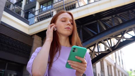 Alegre-chica-pelirroja-bastante-con-los-ojos-cerrados-escuchando-música-usando-auriculares-inalámbricos-en-la-calle-de-la-ciudad