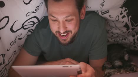 Close-up-of-face-of-a-young-bearded-and-mustashed-man,-using-digital-tablet-under-blanket,-who-is-chatting-and-waves-hand,-in-his-bed,-at-night.-His-yorkshire-terrier-is-lying-next-to-him