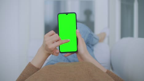 Standpunkt-der-Frau-im-modernen-Zimmer-sitzen-auf-einem-Stuhl-mit-Telefon-mit-grünen-Mock-up-Bildschirm-Chroma-Key-Surfen-Internet-beobachten-Inhalte-Videos-Blogs-Tippen-auf-Center-Screen