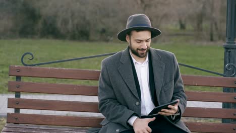 Hipser-creative-business-man-in-hat-using-tablet-pc-computer-and-sitting-on-city-street-bench-in-park-during-break