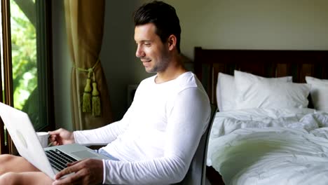 Young-Man-Using-Laptop-Computer-Drink-Coffee-Smiling-Guy-Chatting-Online-In-Bedroom