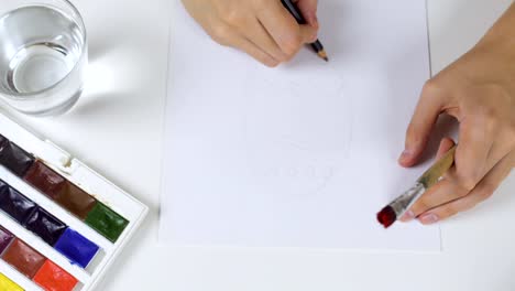 Unrecognizable-woman-draws-a-pencil-sketch-on-paper-Easter-card.-On-the-table-is-a-palette-with-paints-and-a-glass-of-water.