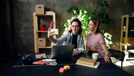 Junge-Unternehmerinnen-machen-Selfie-zusammen-in-modernen-Büro-beim-Sitzen-am-Schreibtisch.-Sie-posiert-mit-lustigen-Gesichtern-und-Lächeln,-dann-Fotos-ansehen.