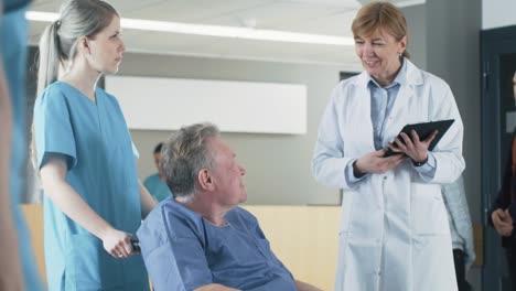 In-the-Hospital-Lobby,-Nurse-Pushes-Elderly-Patient-in-the-Wheelchair,-Doctor-Talks-to-Them-while-Using-Tablet-Computer.-Clean,-New-Hospital-with-Professional-Medical-Personnel.-Slow-Motion.