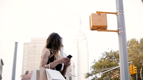 Happy-beautiful-Caucasian-businesswoman-with-shopping-bags-in-sunglasses-using-smartphone-app-near-Empire-State-Building