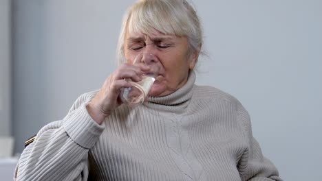 Aging-lady-in-wheelchair-taking-pill-and-drinking-water,-high-blood-pressure