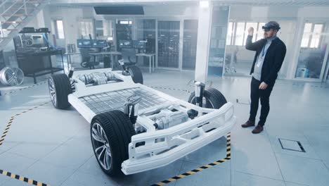 Automotive-Engineer-Working-on-Electric-Car-Chassis-Platform,-Using-Augmented-Reality-Headset.-In-Innovation-Laboratory-Facility-Concept-Vehicle-Frame-Includes-Wheels,-Suspension,-Engine-and-Battery.