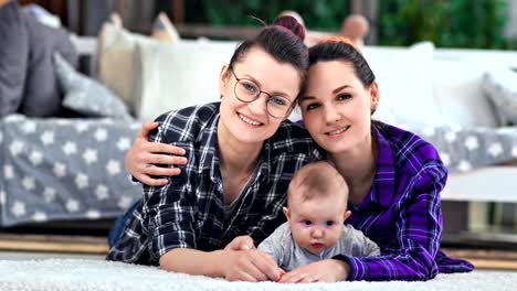 Two-beautiful-hipster-female-parent-posing-with-little-cute-baby-lying-on-floor-looking-at-camera